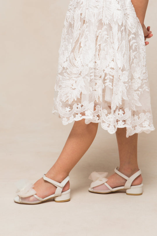 Wearing a white lace dress and comfortable Mini Ivy Bow Heel in White by Ivy City Co—featuring a delicate tulle bow detail—a person stands against a neutral background, with only the lower half of their body visible.