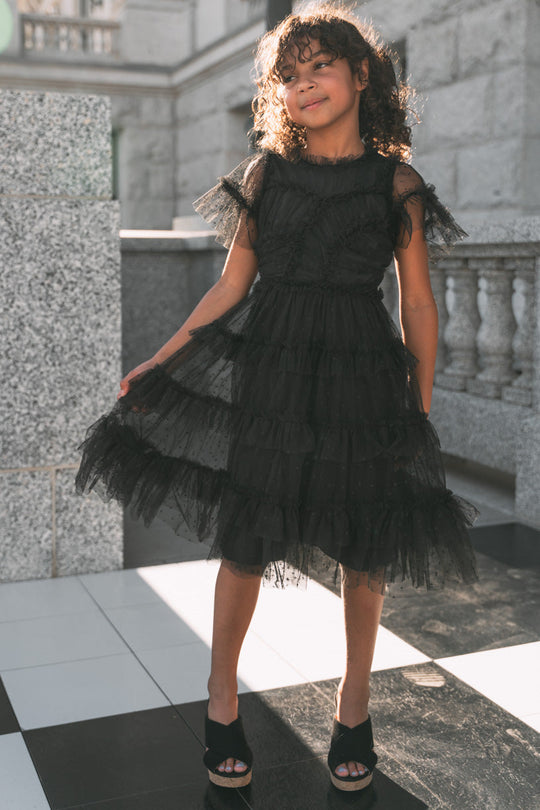 A young girl beams as she stands on a black-and-white tiled floor, sunlight highlighting her curly hair, while she holds out the layered tulle skirt of the Ivy City Co Mini Whimsical Dress in Black.