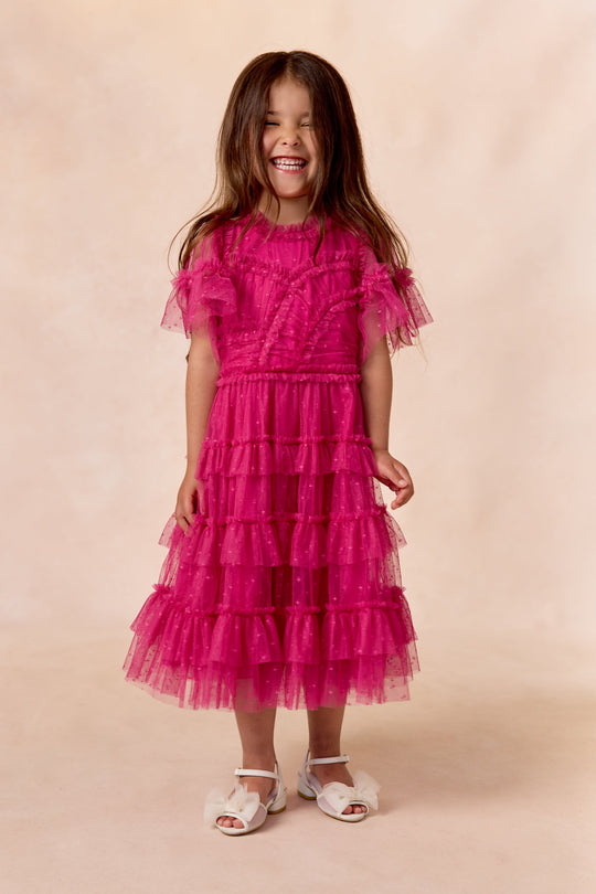 A young girl with long brown hair smiles, wearing the Ivy City Co Mini Whimsical Dress in Fuchsia featuring flutter sleeves and a layered tulle skirt, paired with white sandals, standing against a soft beige background.