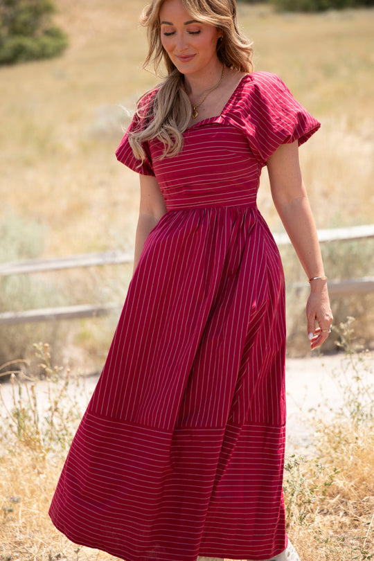 A woman wearing the Ivy City Co Julie Dress in Cranberry Pinstripe, featuring white vertical stripes and puff sleeves, stands outdoors on a sunny day in a dry, grassy area, looking down and smiling slightly.
