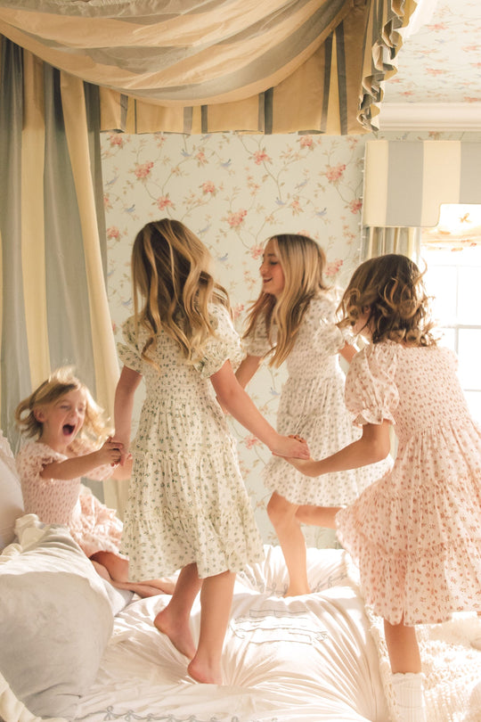 Four young girls in light, floral dresses—including the charming Ivy City Co Mini Madeline Dress in Blue—hold hands and joyfully jump on a bed in a softly lit, vintage-style bedroom with floral wallpaper and draped curtains.