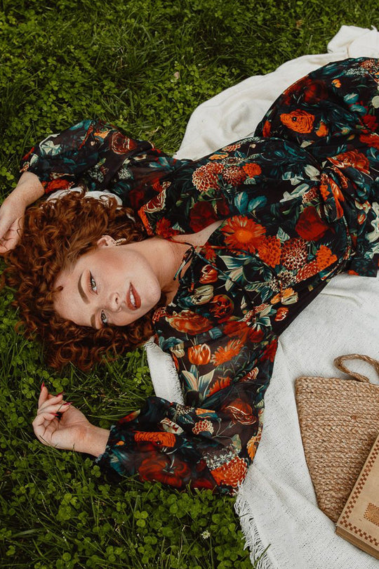 A woman with curly hair reclines on a blanket atop the grass, holding a blade of grass next to a woven bag. She wears the Ivy City Co Imogen Dress in Moody Floral—a vibrant, stylish maxi that’s also maternity friendly—perfect for sun-soaked afternoons.