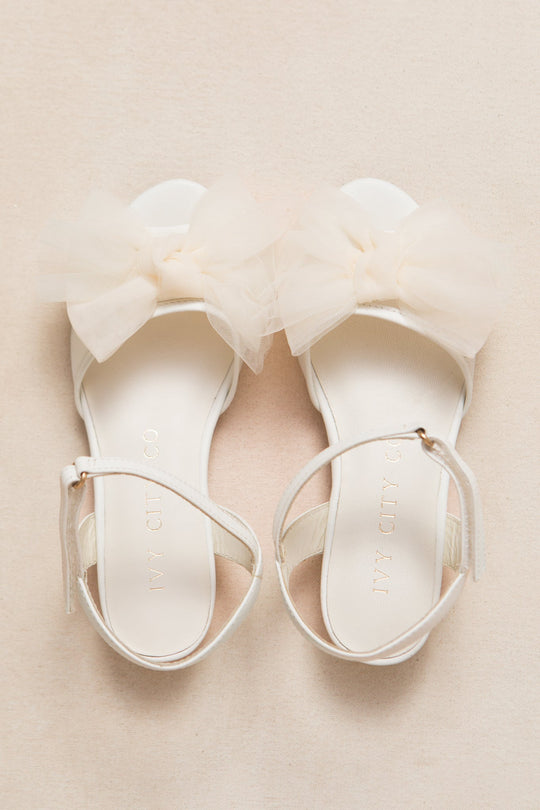 The Mini Ivy Bow Heel in White by Ivy City Co features comfortable white sandals with ankle straps and a large, soft tulle bow detail on the front. Displayed on a light beige surface, these shoes are perfect for any special occasion.