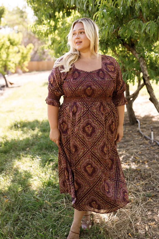 A woman with long blonde hair stands outdoors on grass near leafy trees, enjoying a sunny day while wearing the Ivy City Co Josephine Dress in Nocturne Diamond Rose—a patterned maroon cotton dress featuring a smocked bodice.