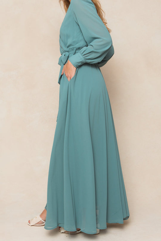 A woman poses sideways with one hand in her pocket, wearing the Ivy City Co Andie Dress in Seaglass Chiffon—a long-sleeved, maxi-length dress with a waist tie. She pairs it with white open-toed shoes against a neutral background.