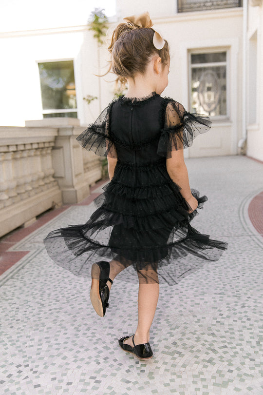 A young girl twirls outdoors on a tiled walkway, her hair tied up with a bow, wearing the Ivy City Co Mini Whimsical Dress in Black—a charming style featuring a black layered tulle skirt—paired with black shoes.