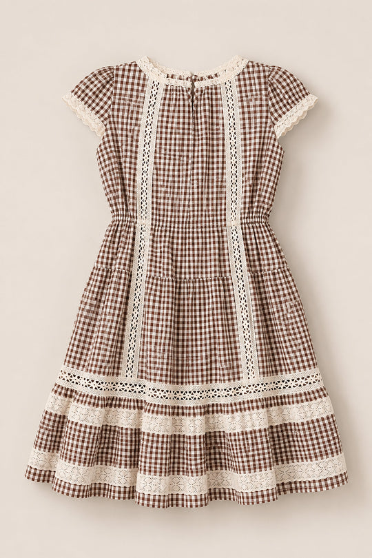 The Mini Palermo Dress in Hickory Gingham by Ivy City Co features a brown and white cotton-blend gingham pattern, short sleeves, and delicate lace trim along the neckline, bodice, sleeves, skirt, and hem. With its slightly flared skirt and vintage cottagecore-inspired design, this girls’ dress brings timeless charm to any occasion.