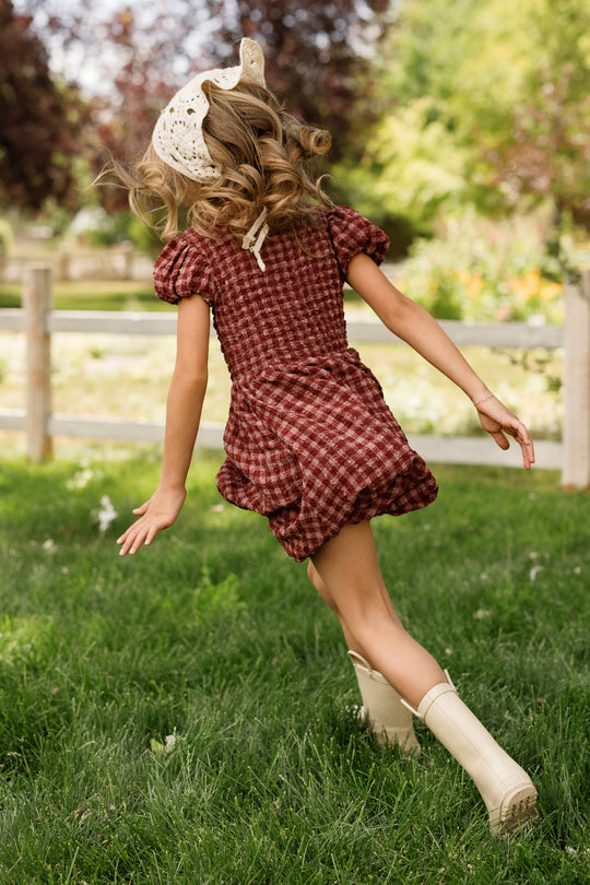 A young girl wearing the Ivy City Co Mini Tori Dress in Cranberry Popcorn Plaid, featuring a smocked bodice and charming puff sleeves, runs across green grass with her curly hair flowing beneath a lace bonnet. Trees and a wooden fence stand in the background.
