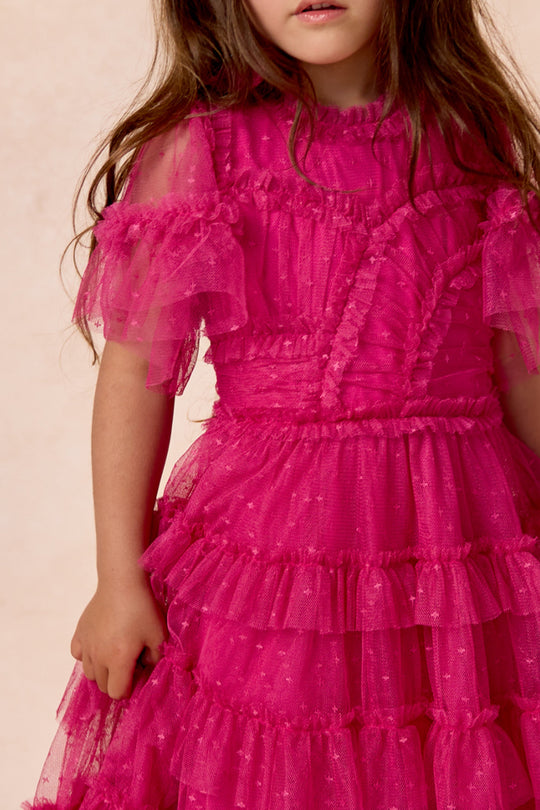A young girl wearing the Ivy City Co Mini Whimsical Dress in Fuchsia, featuring a layered tulle skirt and sheer flutter sleeves, stands against a neutral background.
