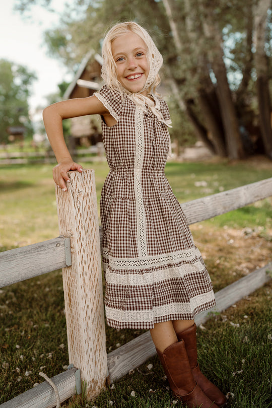 A smiling young girl in the Ivy City Co Mini Palermo Dress in Hickory Gingham, paired with a bonnet and brown boots, leans against a wooden fence outdoors, with trees and a house in the background.