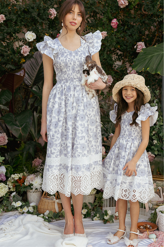 A woman and a girl wear matching Ivy City Co Hattie Dresses in Blue Floral, with lace hems. Outdoors among flowers and greenery, the woman holds a rabbit while the girl sports a straw hat.