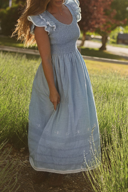 A woman wearing the Ivy City Co Hattie Ankle Length Dress in Embroidered Chambray, featuring a light blue smocked bodice and ruffle sleeves, stands in a sunlit lavender field, gently holding the skirt of her dress. Her face is out of frame.
