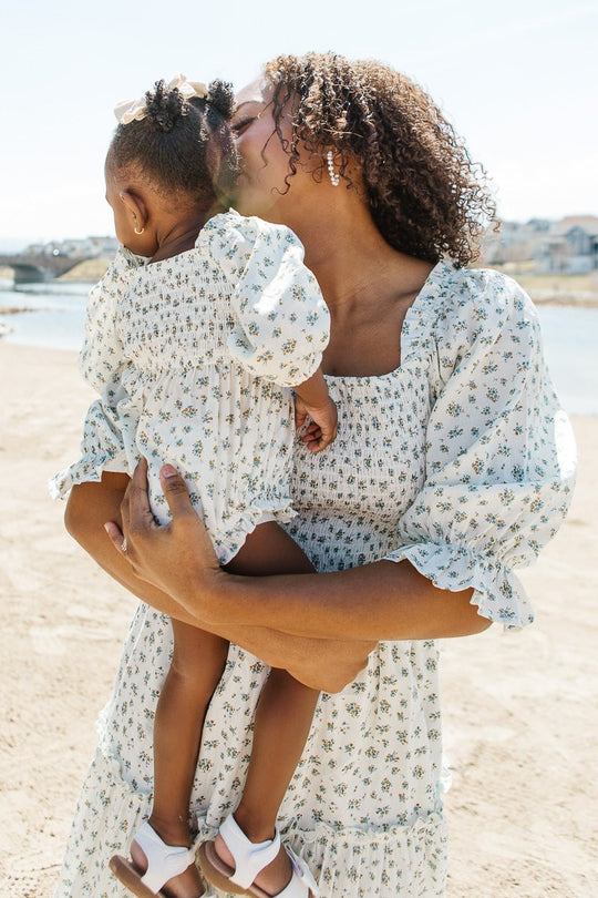 A woman lovingly kisses a toddler on the forehead while holding her. Both are wearing matching blue-floral dresses from Ivy City Co, with the little one in the adorable Baby Madeline Romper in Blue. They stand outdoors near water on a sunny day.