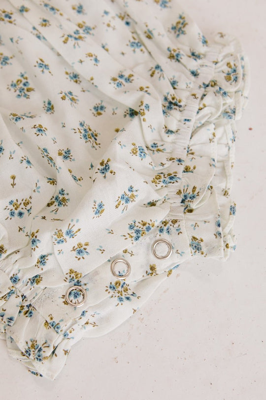 Close-up of the Ivy City Co Baby Madeline Romper in Blue, showcasing white fabric adorned with a blue and yellow floral pattern, metal snap buttons, and ruffled edges; displayed against a light, textured background.