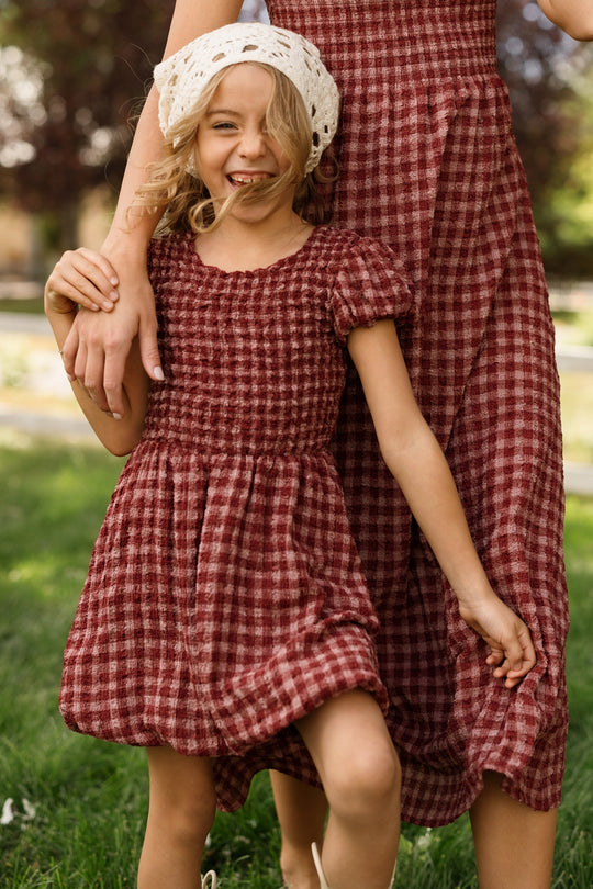 A young girl wearing the Ivy City Co Mini Tori Dress in Cranberry Popcorn Plaid with puff sleeves and a white crochet hat smiles as she holds hands with an adult in a coordinating dress, both standing on grass outdoors.