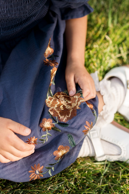 A child sits on the grass wearing the Ivy City Co Mini Teagan Dress in Navy Embroidered Fall Floral, which features a smocked bodice and embroidered flowers, along with white socks and white shoes. Only the child’s hands and part of their legs are visible.