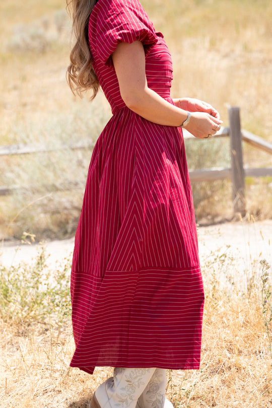 A woman stands outdoors on a sunny day, hands together and facing sideways, wearing the Ivy City Co Julie Dress in Cranberry Pinstripe. The dress features a smocked bodice and puff sleeves, adding charm to her look. A wooden fence and dry grass are visible in the background.
