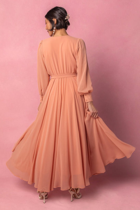 A woman wearing the Ivy City Co Andie Dress in Apricot Crush stands against a pink background, facing away. She holds part of her maxi dress, with her hair styled up and adorned with a decorative clip, and wears high-heeled sandals.