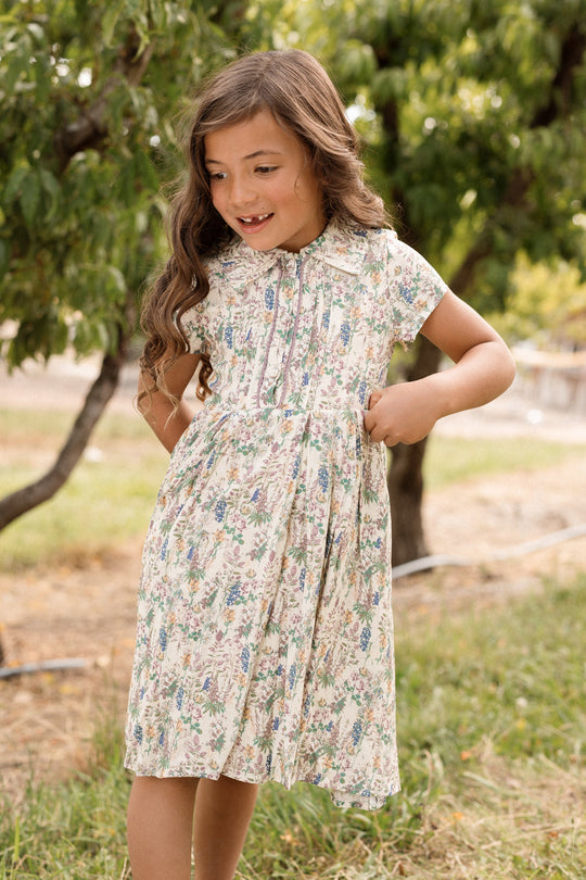 A young girl with long brown hair stands outside on the grass near trees, smiling and looking to the side with one hand on her hip and the other holding her Mini Amber Dress in Autumn Bouquet by Ivy City Co.