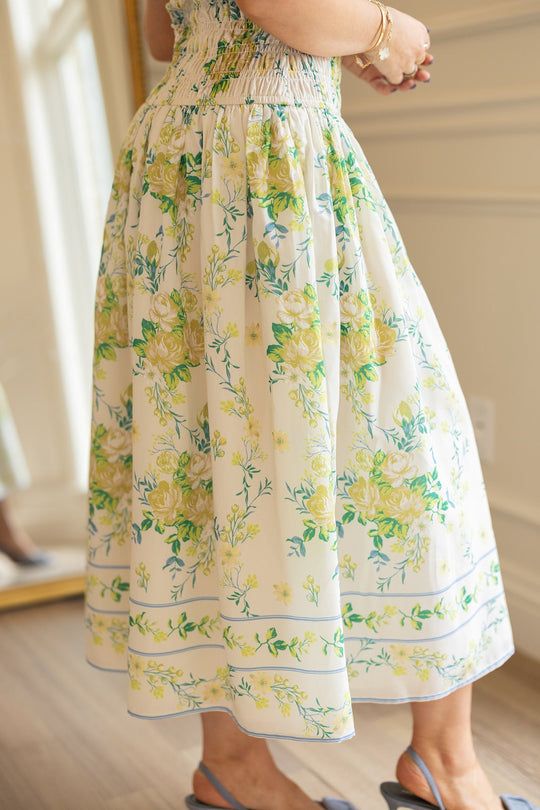 A person stands indoors wearing the Ivy City Co Jennifer Dress in Garden Tea Party Floral—a yellow and green midi dress with a gathered waist made from lightweight fabric—paired with blue sandals. Their hands are clasped, displaying gold bracelets, while a mirror and paneled wall appear in the background.