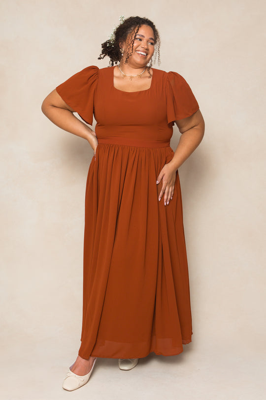 A woman in the Ivy City Co Abbie Dress in Amber Chiffon, a rust-colored maxi with short puff sleeves, smiles against a beige background. She has curly hair, a necklace, and cream flats.