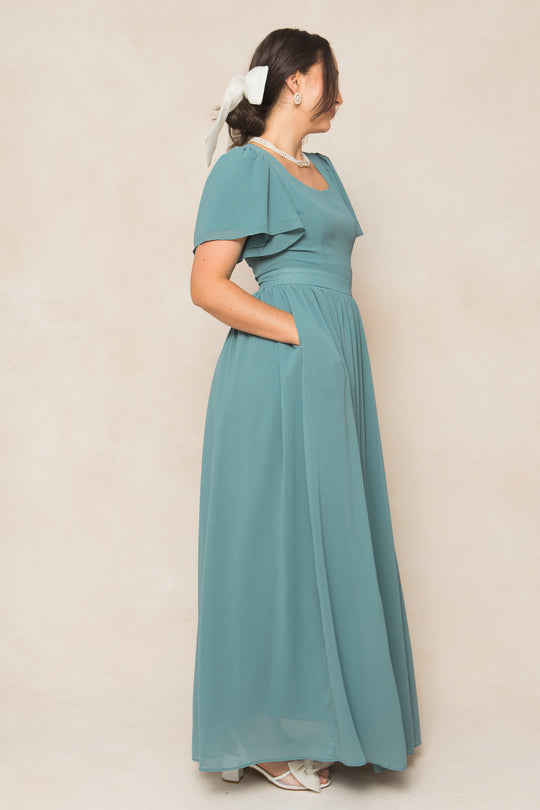 A woman stands in profile against a beige backdrop wearing the Ivy City Co Abbie Dress in Seaglass Chiffon with pockets, styled with white open-toe heels, a pearl necklace, earrings, and a large white bow—ideal for fall bridesmaids.