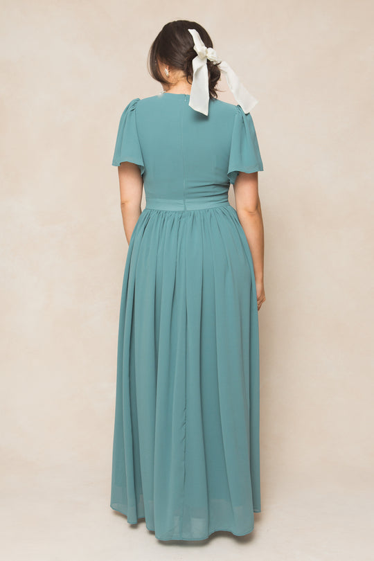 A woman with dark hair in a low ponytail tied with a white ribbon wears the Ivy City Co Abbie Dress in Seaglass Chiffon, a floor-length, short-sleeved gown, standing with her back to the camera against a beige background.