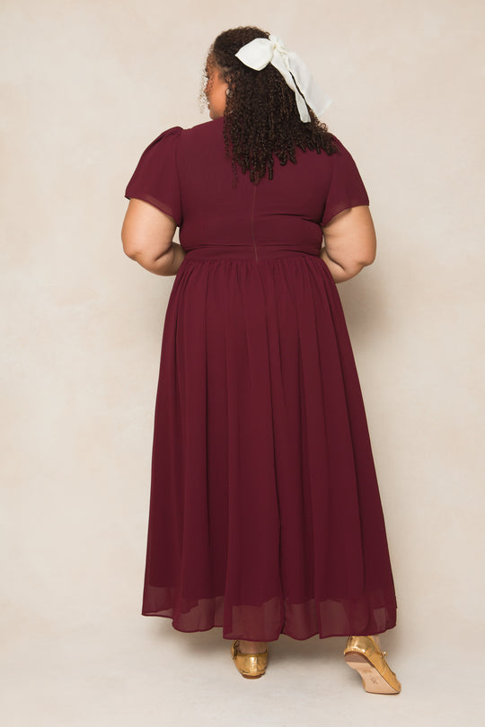 A woman stands with her back to the camera in the Ivy City Co Abbie Dress in Wine Chiffon, featuring curly hair tied with a large white ribbon and gold shoes—a perfect fall bridesmaid look against a neutral background.
