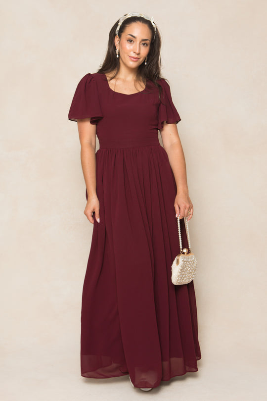 A woman in the Ivy City Co Abbie Dress in Wine Chiffon, a short-sleeve, floor-length bridesmaid dress, stands against a light background holding a beaded purse and wearing a headband with her hair down and a neutral expression.