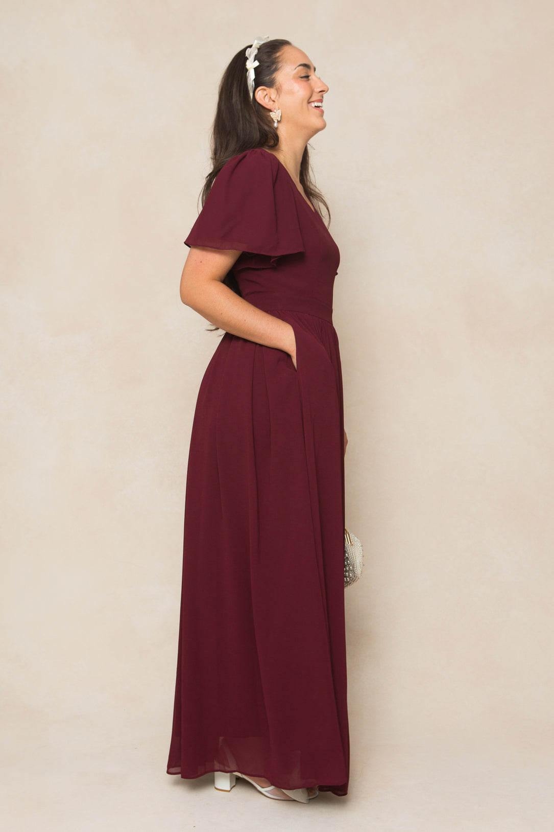 A woman with long dark hair smiles in profile, wearing the Ivy City Co Abbie Dress in Wine Chiffon and white heels. She accessorizes with a white headband and beaded clutch—perfect as a fall bridesmaid dress.