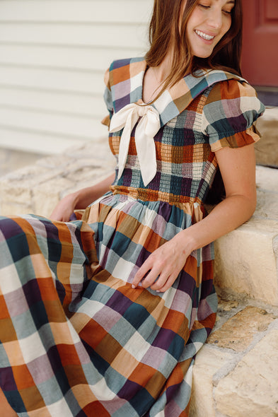 A woman sits on stone steps, smiling and looking down, wearing the Ivy City Co Alice Dress in Maize Gingham with puff sleeves and a large white bow at the collar.