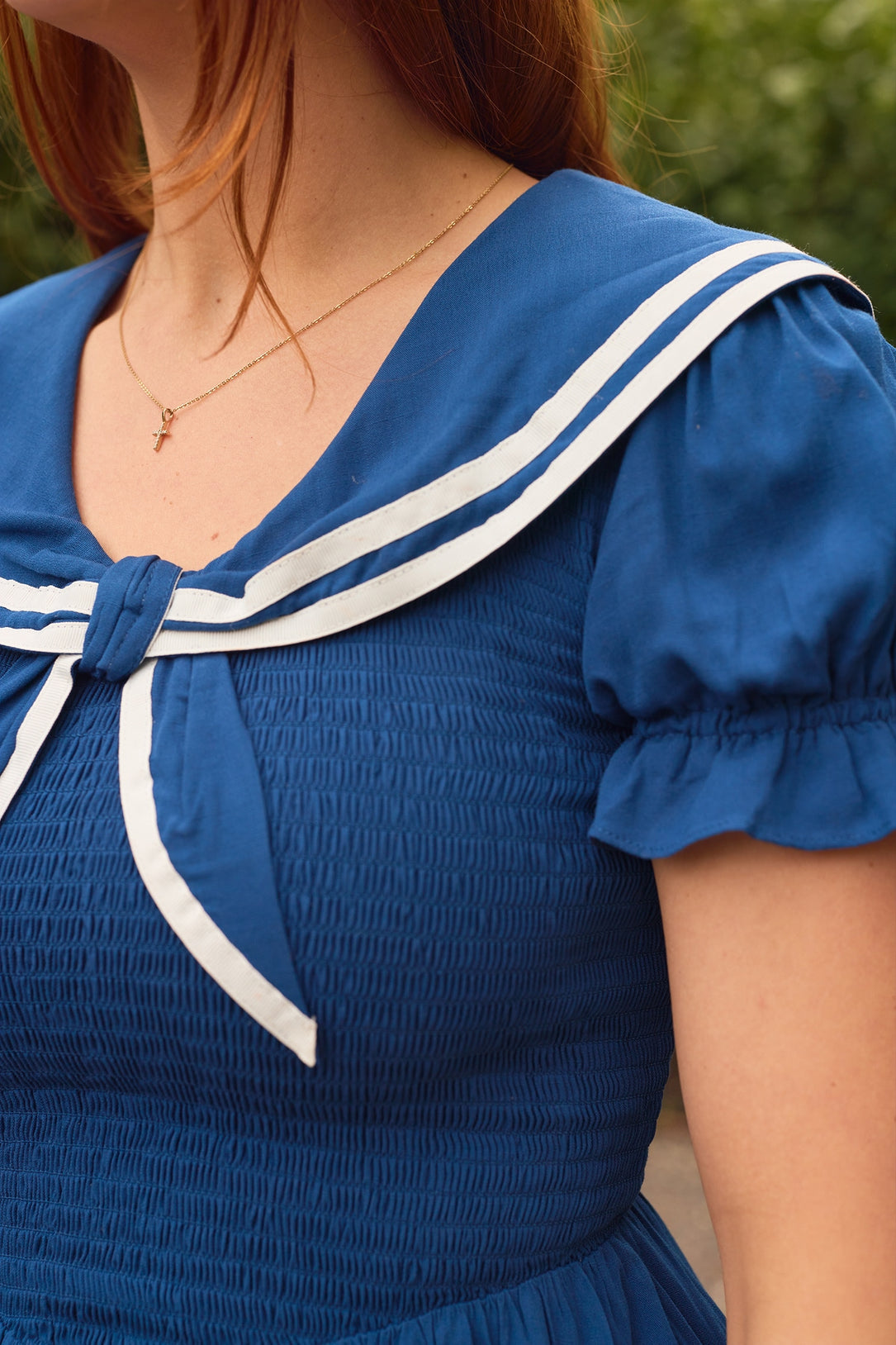 A person wears the Ivy City Co Alice Dress in Sailor Navy, featuring a white-trimmed sailor collar and puffed sleeves. The smocked, maternity-friendly dress is styled with long hair and a delicate necklace against blurred greenery.