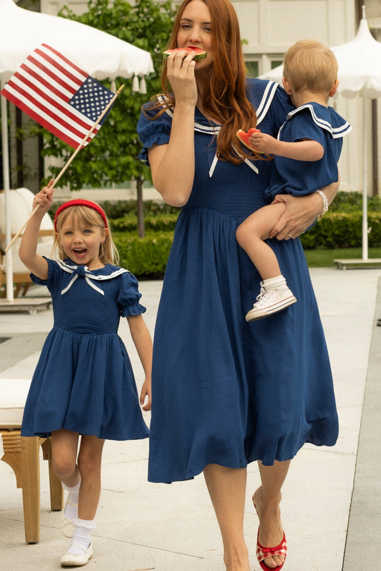 A woman and two children wear Ivy City Co’s Alice Dress in Sailor Navy. The mom holds a toddler and eats watermelon while the older child waves an American flag, showing how this maternity-friendly dress is perfect for sunny summer outings.