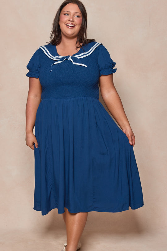 A woman smiles and holds one side of her Ivy City Co Alice Dress in Sailor Navy, featuring short sleeves and a knee-length skirt, against a beige background.