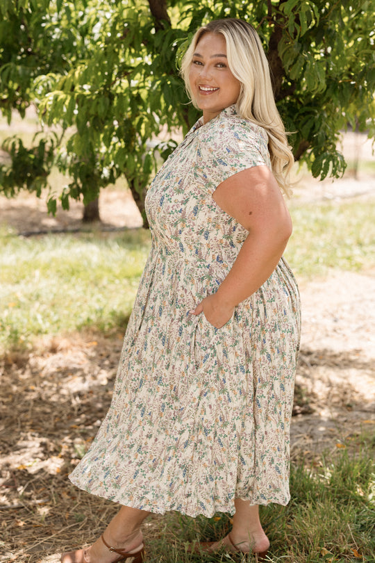 A woman with long blonde hair stands outdoors near a leafy tree on a sunny day, smiling as she wears the Ivy City Co Amber Dress in Autumn Bouquet—a nursing-friendly, tea-length floral dress—paired with sandals.