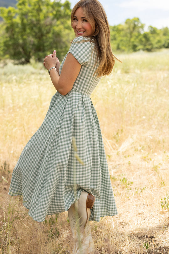 A woman wearing the Ivy City Co Amber Dress in Sage Gingham stands in a sunlit field, surrounded by tall grass and greenery, turning slightly to look over her shoulder with trees in the background.