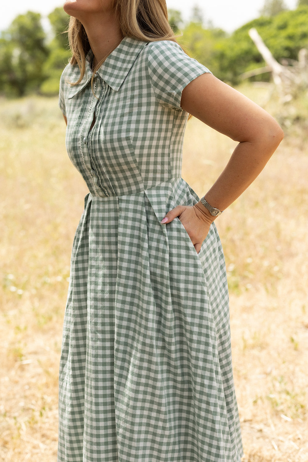 A woman stands outdoors with hands in her pockets, wearing the Ivy City Co Amber Dress in Sage Gingham. The short-sleeve dress contrasts against a grassy background with blurred trees. Her face is not shown.