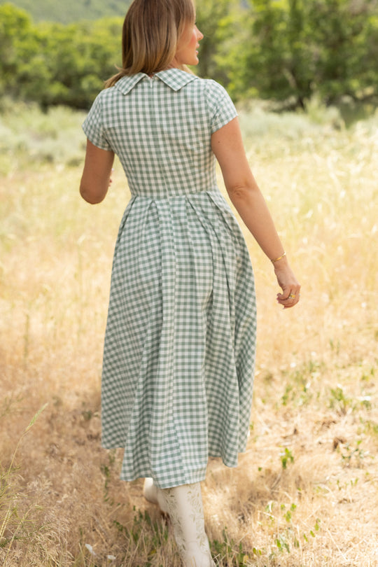 A woman wearing the Ivy City Co Amber Dress in Sage Gingham, featuring a pleated skirt, walks through a sunlit field of dry grass with green trees and rolling hills, her back to the camera.