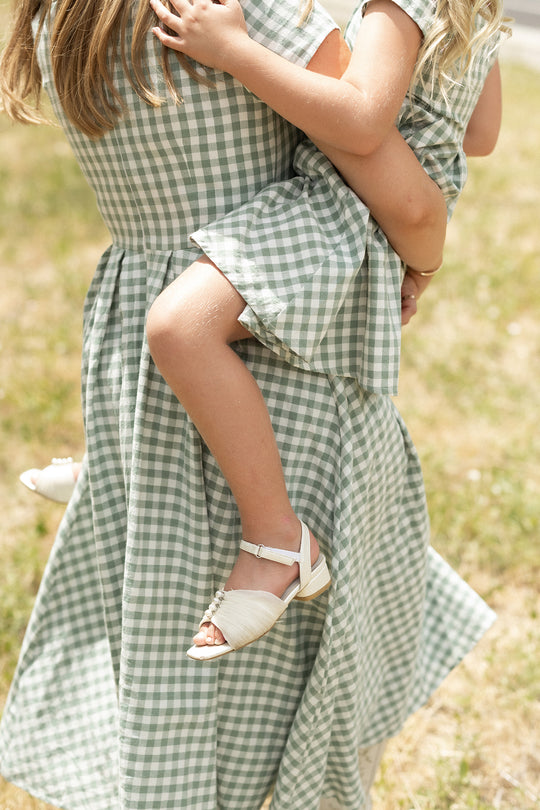 A woman in the Ivy City Co Amber Dress in Sage Gingham holds a young girl in a matching cotton midi dress and white sandals, standing on grass under bright daylight. Their faces are not shown.