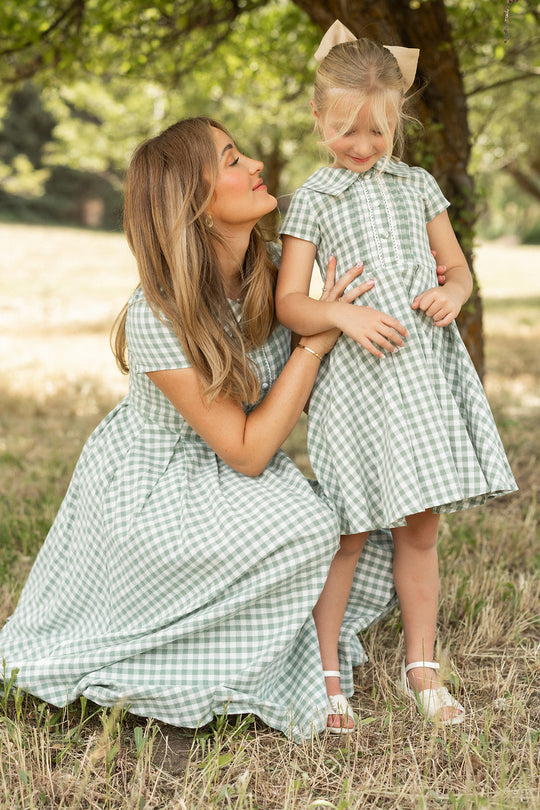 A woman and a young girl stand outdoors under a tree, both wearing the Ivy City Co Amber Dress in Sage Gingham. The woman smiles as she gently holds the girl's arm, while the girl shyly smiles in her matching cotton midi dress.
