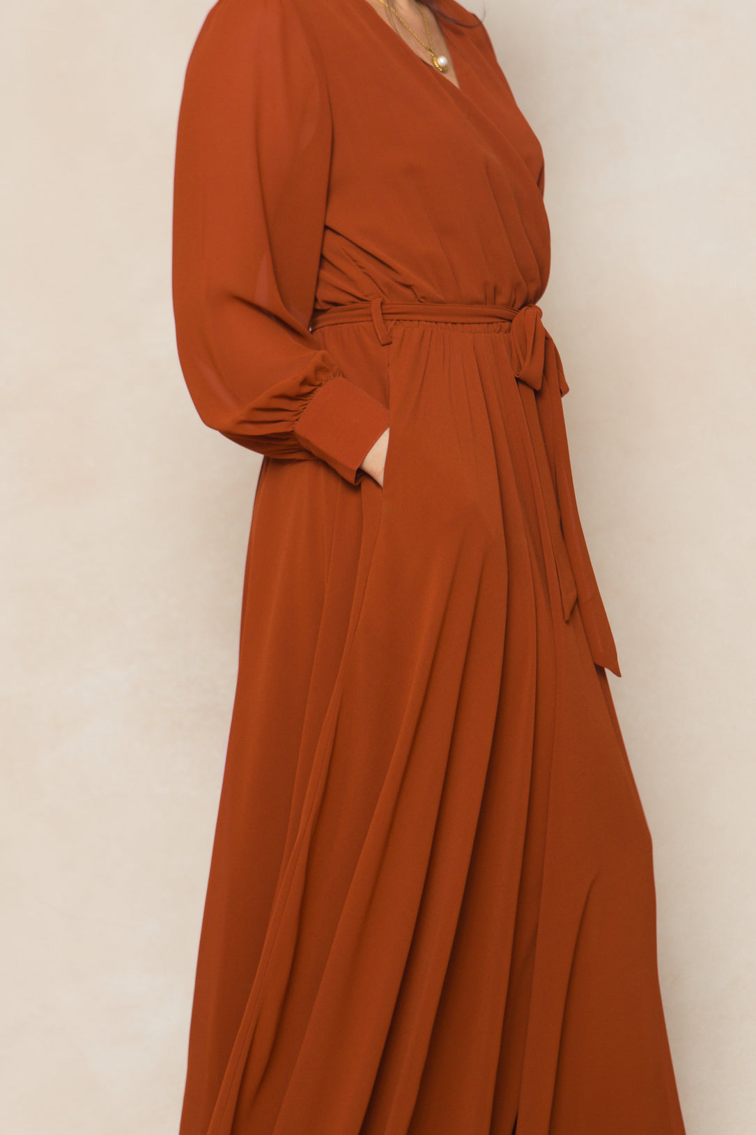 A woman models Ivy City Co's Andie Dress in Amber Chiffon—a rust-hued maxi with long sleeves, waist tie, and side pockets—against a light beige backdrop, offering an elegant option for fall bridesmaids.