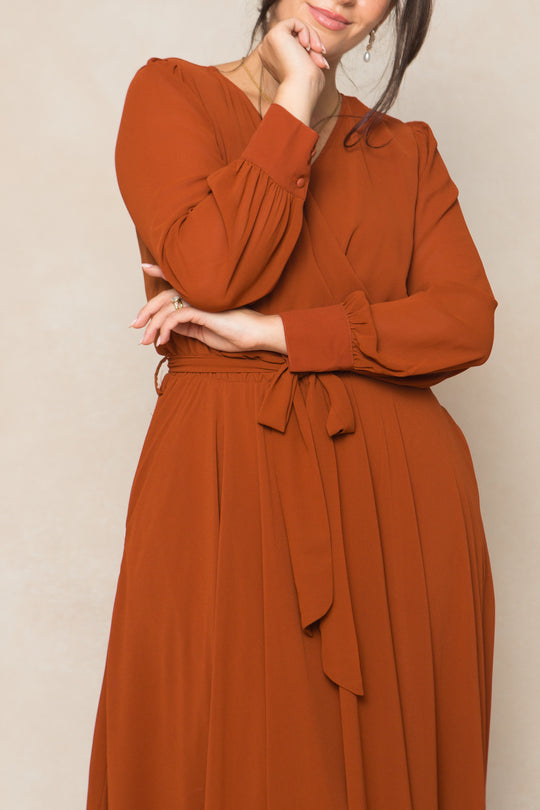 A woman with dark hair pulled back models the Ivy City Co Andie Dress in Amber Chiffon—a long-sleeved maxi dress with a waist tie—as she poses with one arm crossed and the other hand touching her chin, her face partially visible.