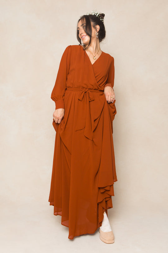 Woman wearing the Ivy City Co Andie Dress in Amber Chiffon—a long maxi dress with sleeves and waist tie—stands against a beige backdrop, smiling, with a floral headpiece and holding her fall bridesmaid dress.