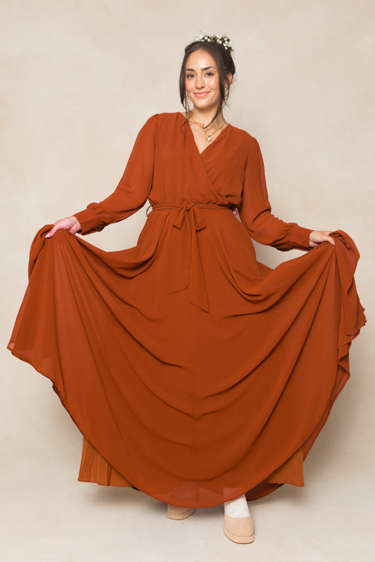 A woman models the Ivy City Co Andie Dress in Amber Chiffon, a long maxi with waist tie, against a beige background. She holds out her skirt and wears her light brown hair up, accessorized with a floral piece.
