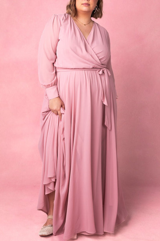 A person wearing the Ivy City Co Andie Dress in Blush, a long maxi dress featuring long sleeves and a waist tie, stands on a pink background. They pair the dress with textured slip-on shoes.