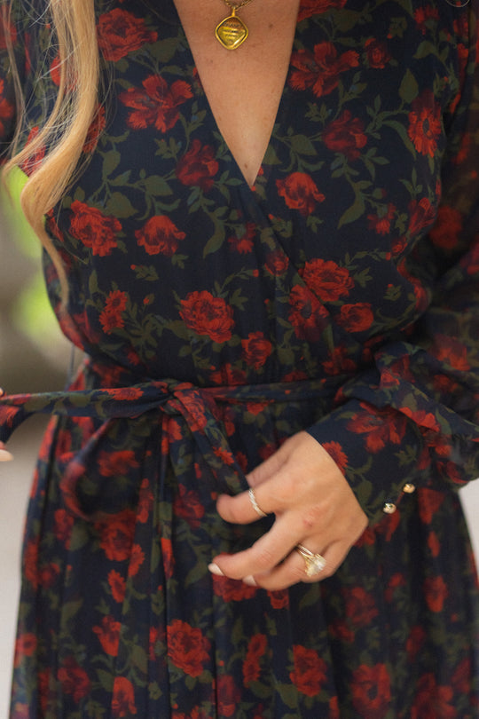 Wearing the Andie Dress in Scarlet Blooms by Ivy City Co, a person with long hair adjusts the waist tie of the dark floral maxi dress adorned with vivid red blooms. They accessorize with rings and a gold necklace featuring a round pendant.