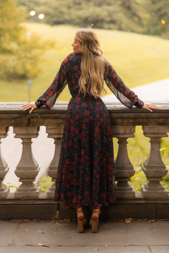 A woman with long blonde hair wears the Ivy City Co Andie Dress in Scarlet Blooms—a navy fall floral maxi featuring red roses and long sheer balloon sleeves—paired with tan platform heels as she leans on a stone balustrade overlooking a green park.
