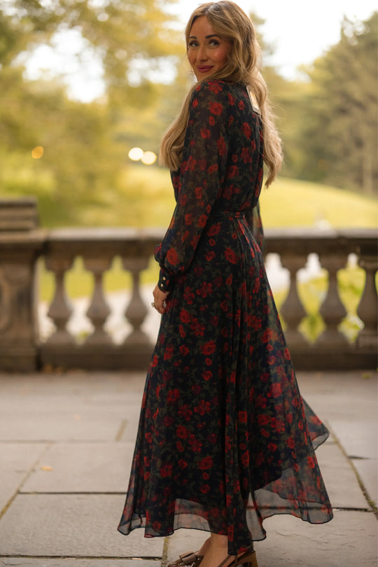 A woman in the Andie Dress in Scarlet Blooms by Ivy City Co—a long, sheer black maxi dress adorned with red floral prints—stands on a stone terrace, turning to look back over her shoulder. Green trees and grass are softly blurred in the background.