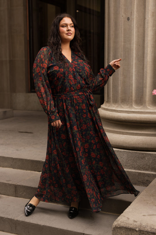 Wearing the Ivy City Co Andie Dress in Scarlet Blooms—featuring elegant black and red fall floral patterns—a woman stands confidently on stone steps beside a grand column, exuding stylish sophistication.