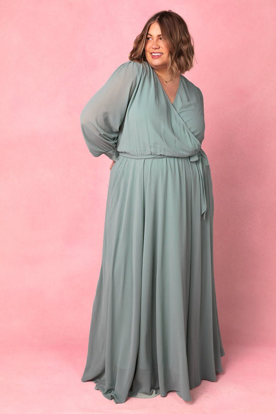 A smiling woman with shoulder-length hair wears the Ivy City Co Andie Dress in Sage, a flowing bridesmaid dress with long sleeves, standing against a soft pink background.